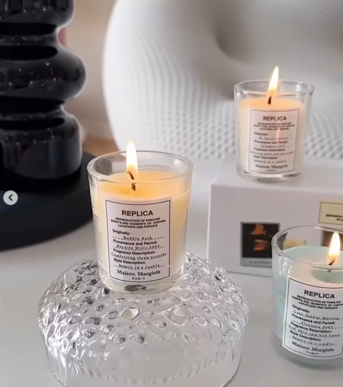 These are Maison Margiela Replica scented candles, featuring glass jars with minimalist white cotton labels reminiscent of their fragrance bottles.